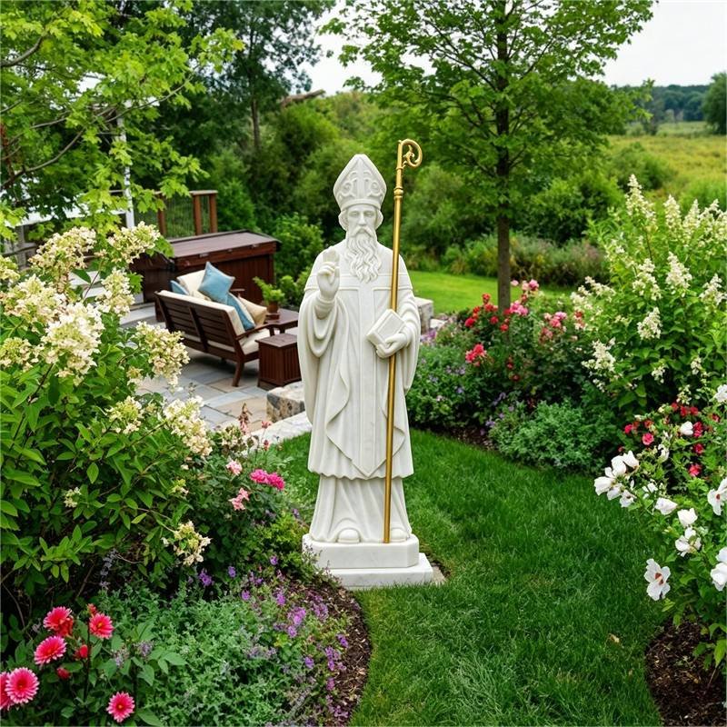 White marble statue of Saint Patrick with a golden crozier, standing in a lush flower garden.