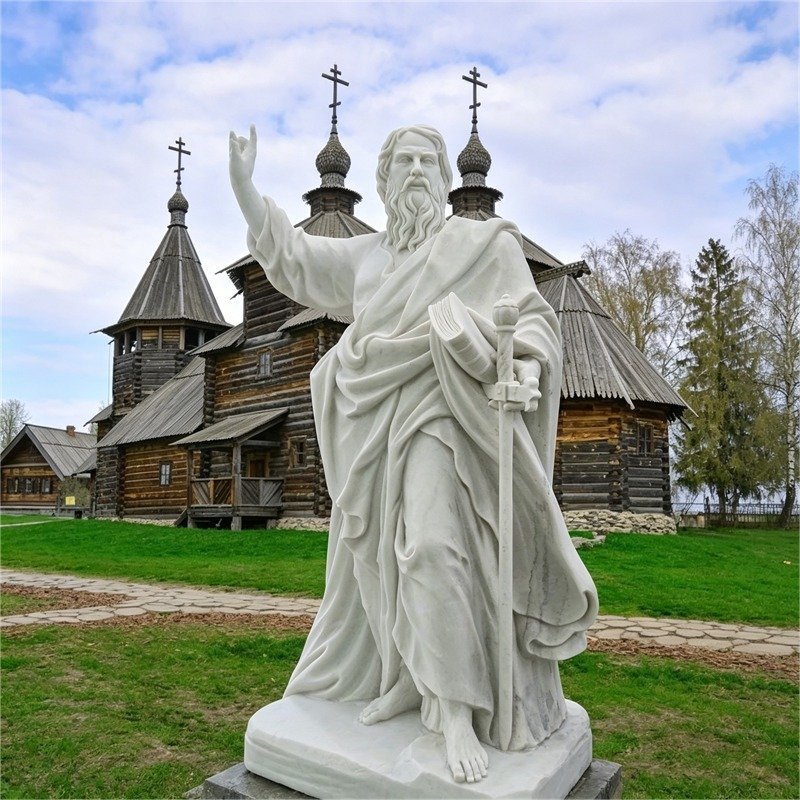 White marble statue of Saint Paul with a sword and book, standing in front of a traditional Russian wooden church.