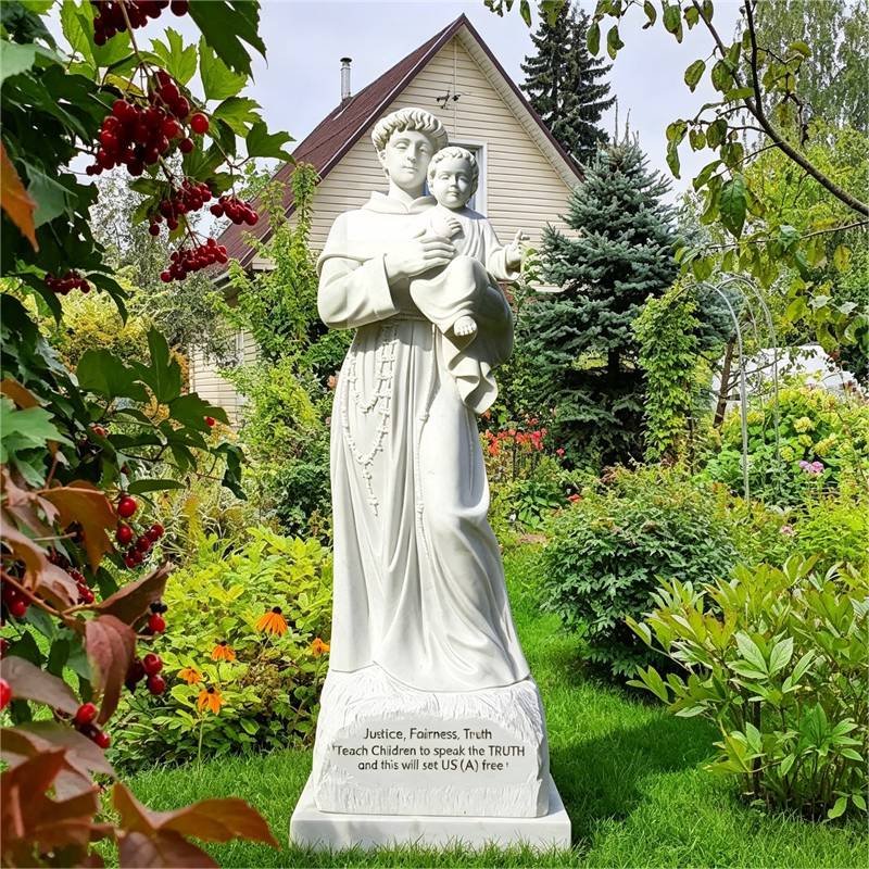 White marble statue of Saint Anthony holding Baby Jesus, set in a lush green garden with a house in the background.