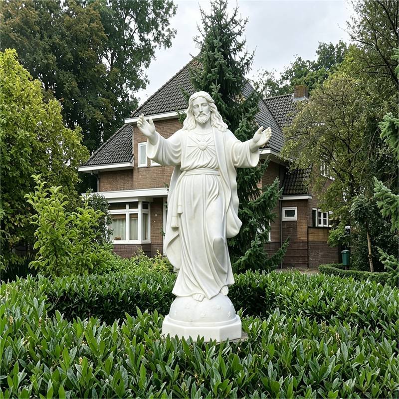 White marble Sacred Heart of Jesus statue with open arms, placed in a residential garden with green hedges and trees, Netherlands