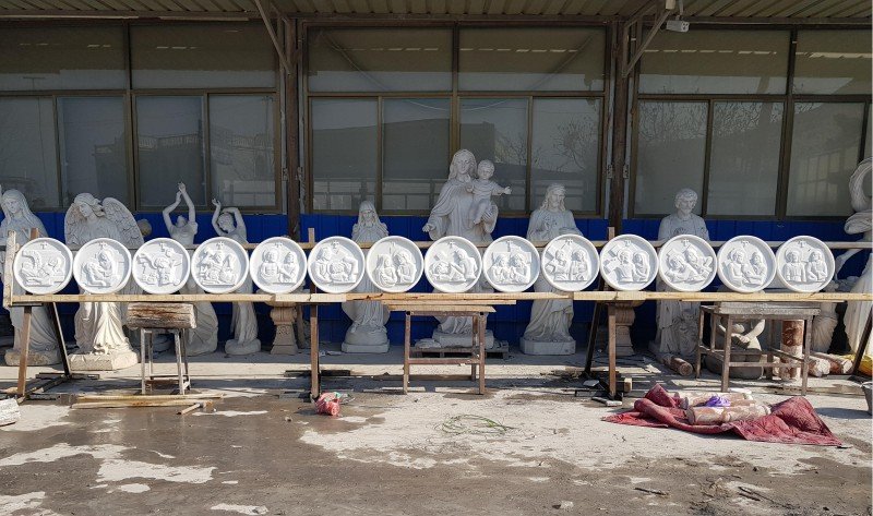 Fourteen white circular Stations of the Cross reliefs displayed on a rack in the workshop